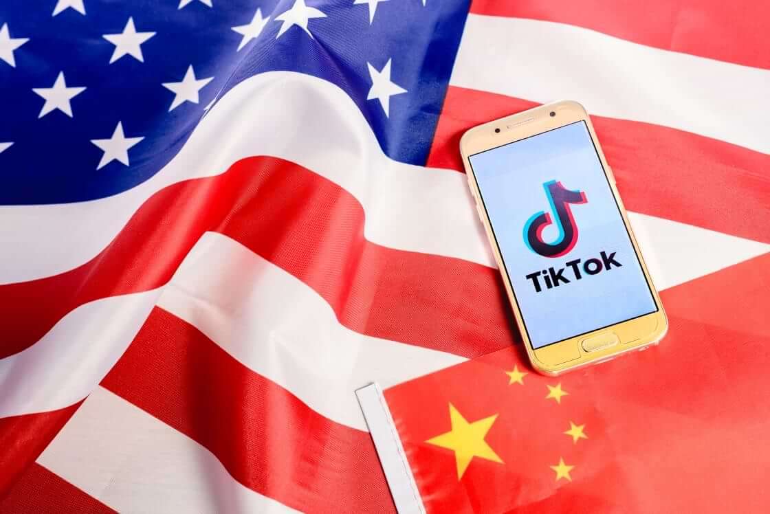 A smartphone displaying the TiTok logo sits atop a Chinese flag and an American flag, illustrative of the long-discussed TikTok ban that may occur in the US. © By Joaquin Corbalan/stock.adobe.com