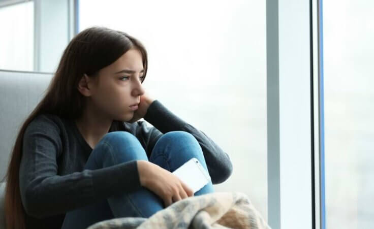 Depressed teen looks out window holding a phone