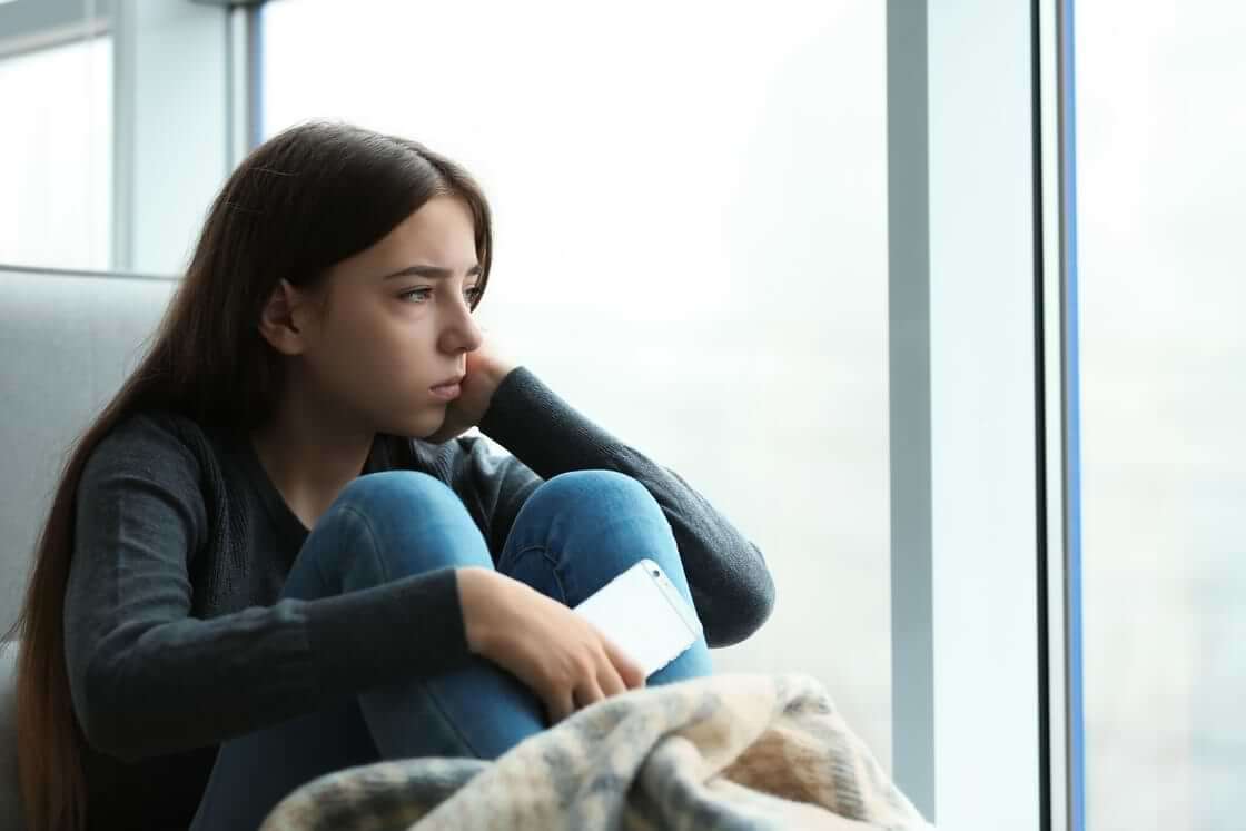 Depressed teen looks out window holding a phone