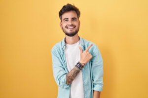 Un joven hispano sonriente con tatuajes se para frente a un fondo amarillo, apuntando hacia arriba y hacia la izquierda con la mano derecha y el dedo índice. © By Krakenimages.com/stock.adobe.com