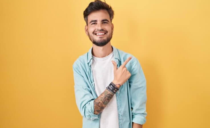 Un joven hispano sonriente con tatuajes se para frente a un fondo amarillo, apuntando hacia arriba y hacia la izquierda con la mano derecha y el dedo índice. © By Krakenimages.com/stock.adobe.com