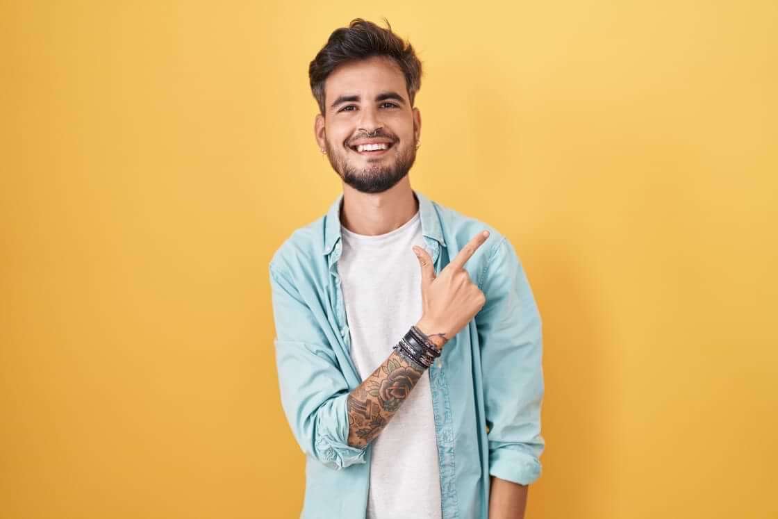Un joven hispano sonriente con tatuajes se para frente a un fondo amarillo, apuntando hacia arriba y hacia la izquierda con la mano derecha y el dedo índice. © By Krakenimages.com/stock.adobe.com