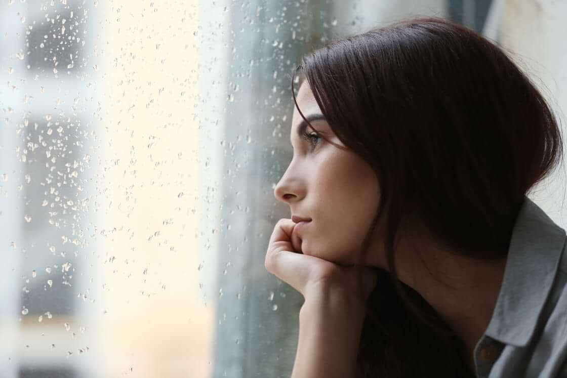 A lonely young woman stares out of a rainy window. © By Africa Studio/stock.adobe.com. Per the US Surgeon General's office, loneliness “can increase the risk of premature death to levels comparable to smoking daily.”
