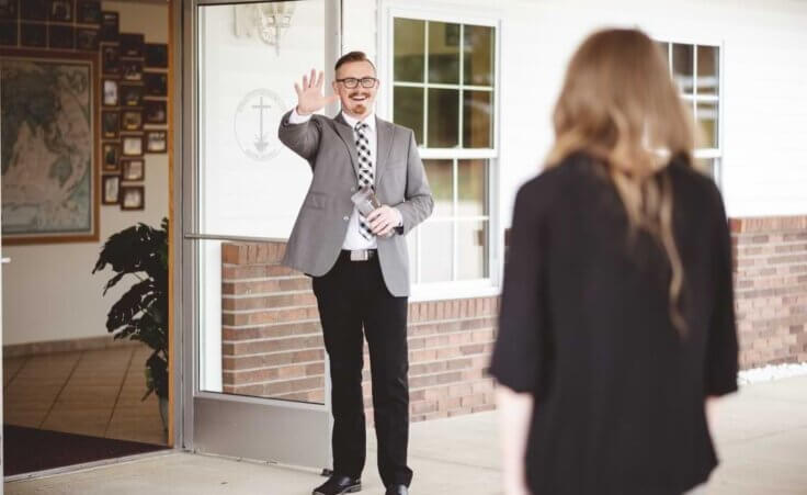 While standing outside a church's open door, a pastor greets a woman. © By Ben White/Wirestock/stock.adobe.com