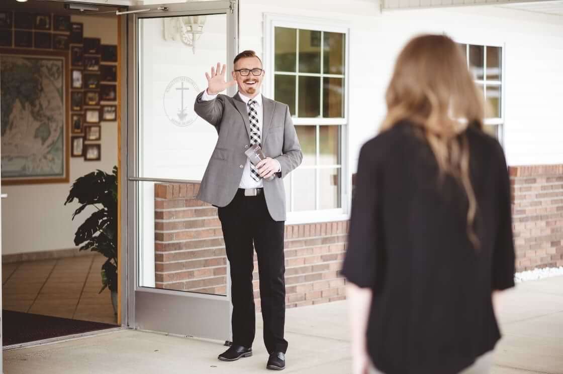 While standing outside a church's open door, a pastor greets a woman. © By Ben White/Wirestock/stock.adobe.com