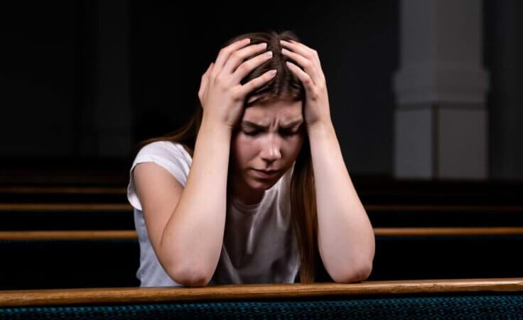 A young woman in a white T-shirt holds her head in her hands while sitting in a church pew, an illustration of having experienced trauma. © By lunarts_studio/stock.adobe.com