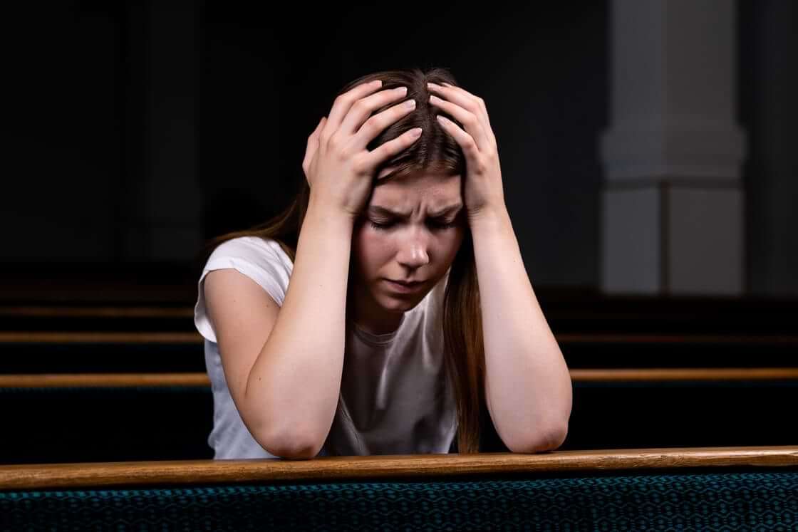 A young woman in a white T-shirt holds her head in her hands while sitting in a church pew, an illustration of having experienced trauma. © By lunarts_studio/stock.adobe.com