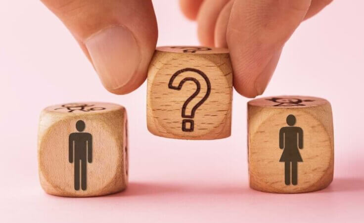 Fingers hold a wooden block emblazoned with a question mark between two other wooden blocks, one depicting a man and one a woman, an illustration of the question: Is cross-dressing a sin? © By Sergey Chayko/stock.adobe.com