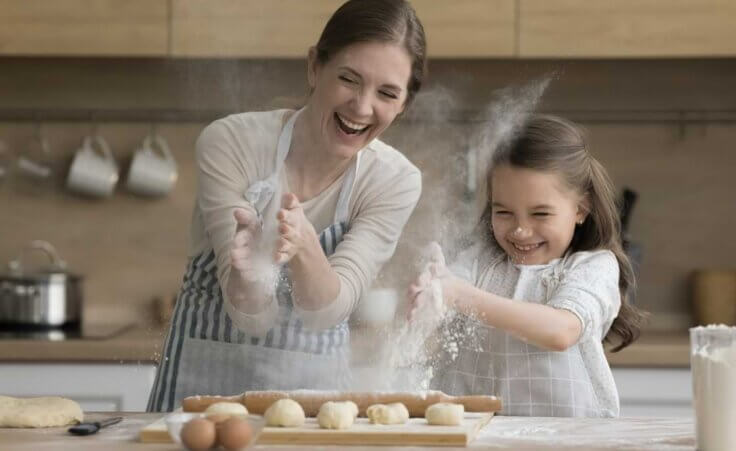 A mother and daughter clap their hands together, sending flour into the air, while making bread. © By fizkes/stock.adobe.com