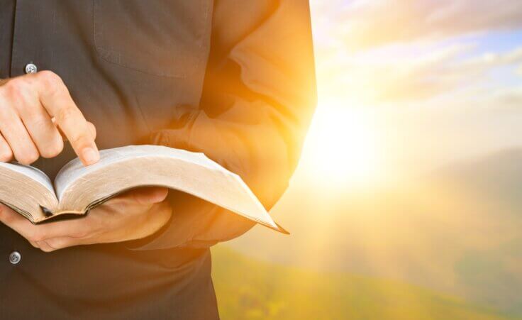 In front of a bright, sunlit background, a man holds an open Bible in his palm while pointing down to a verse with his index finger. © By BillionPhotos.com/stock.adobe.com