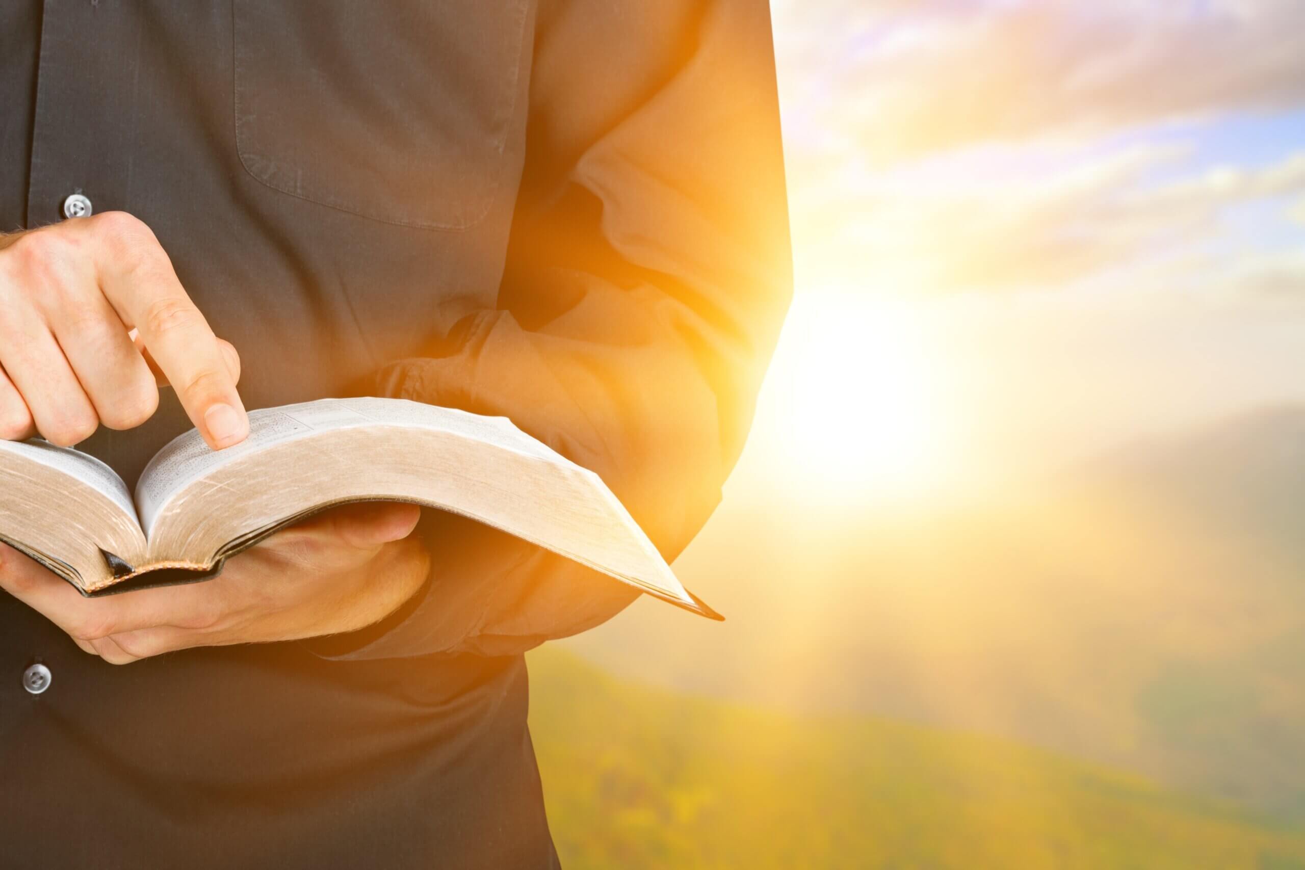In front of a bright, sunlit background, a man holds an open Bible in his palm while pointing down to a verse with his index finger. © By BillionPhotos.com/stock.adobe.com