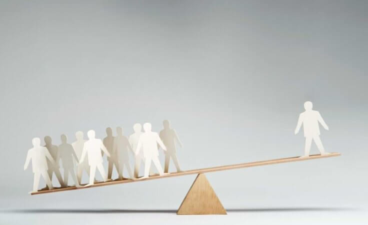 A lone paper figure in the shape of a man stands on the high end of a seesaw with the opposite end supporting ten similar cutouts. © By pogonici/stock.adobe.com