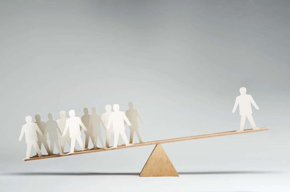 A lone paper figure in the shape of a man stands on the high end of a seesaw with the opposite end supporting ten similar cutouts. © By pogonici/stock.adobe.com