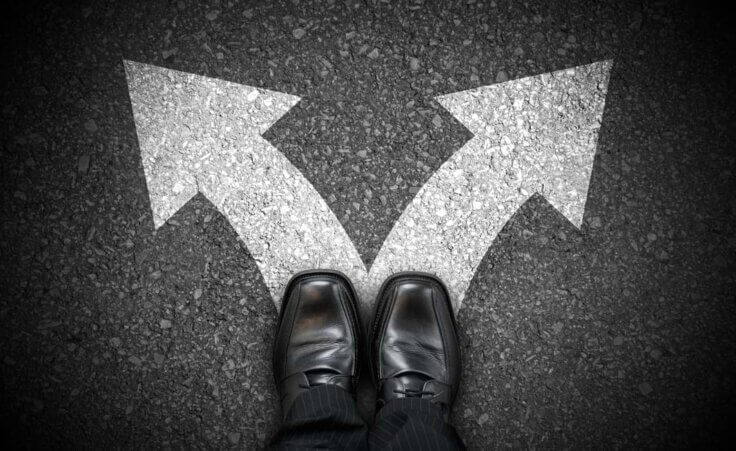 Black business shoes stand on two white arrows pointing away from each other on an asphalt road. © By PX Media/stock.adobe.com