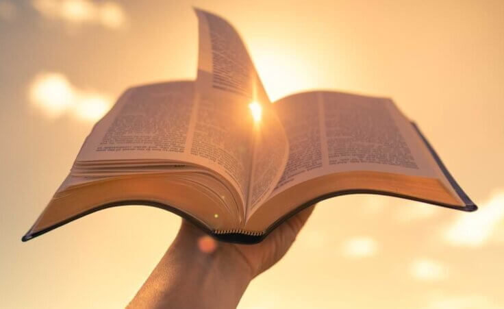 A hand holds an open Bible against a sunlit sky. © By kieferpix/stock.adobe.com