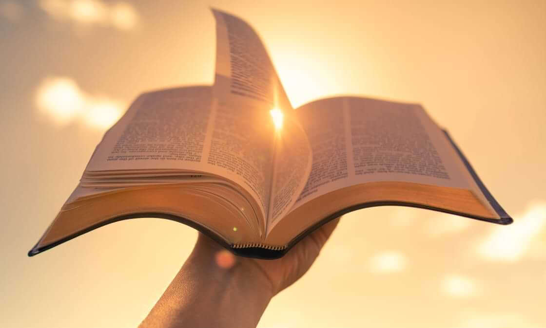 A hand holds an open Bible against a sunlit sky. © By kieferpix/stock.adobe.com