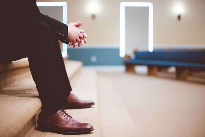 A pastor sits on the front steps of his church pulpit, his hands clasped in front of him, with pews in the background. Photo by Ben White on Unsplash