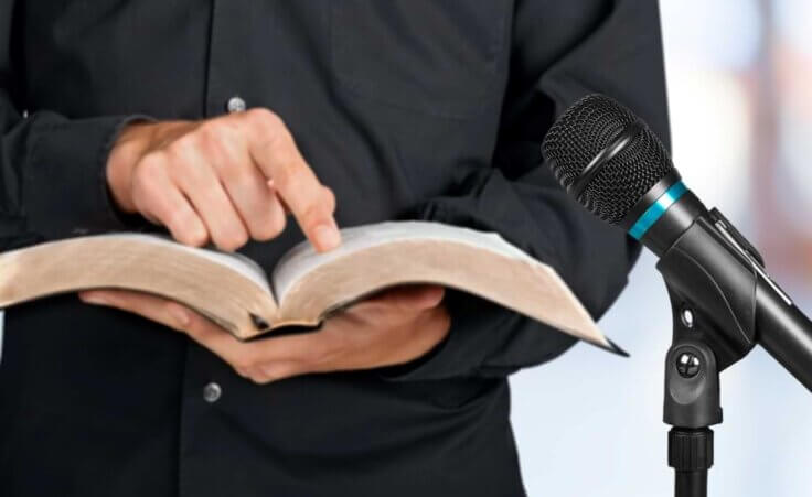 A preacher clad in black stands next to a microphone while holding an open Bible in his left palm, his index finger on his right hand pointing to a passage of Scripture. © By BillionPhotos.com/stock.adobe.com. In Titus 2:12, Paul writes that Christians are to "declare these things; exhort and rebuke with all authority."