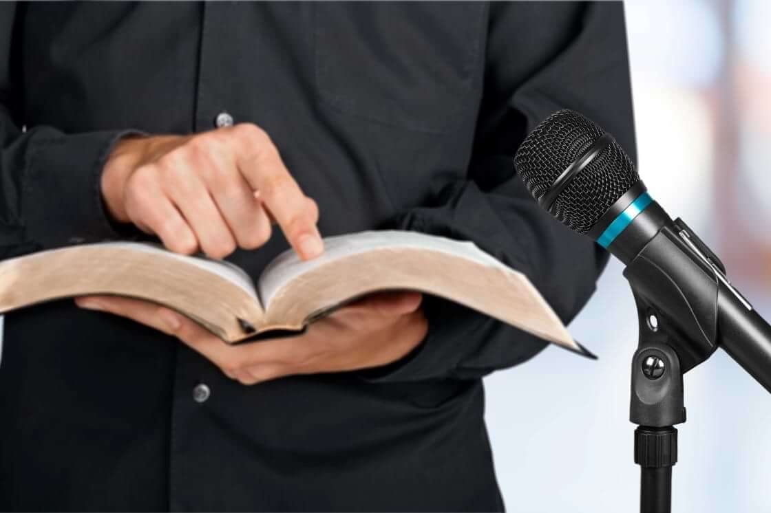 A preacher clad in black stands next to a microphone while holding an open Bible in his left palm, his index finger on his right hand pointing to a passage of Scripture. © By BillionPhotos.com/stock.adobe.com. In Titus 2:12, Paul writes that Christians are to "declare these things; exhort and rebuke with all authority."