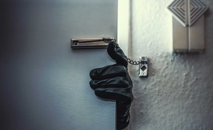 Closeup of a black-gloved hand opening a blue door from the outside, a chain-latched lock still holding the door closed, indicating a thief breaking in © By AA+W/stock.adobe.com. Marjorie Perkins, 87, fought off a teenage intruder who’d struck her. Then, when the teen said he was “awfully hungry,” she fed him—and then called 911.
