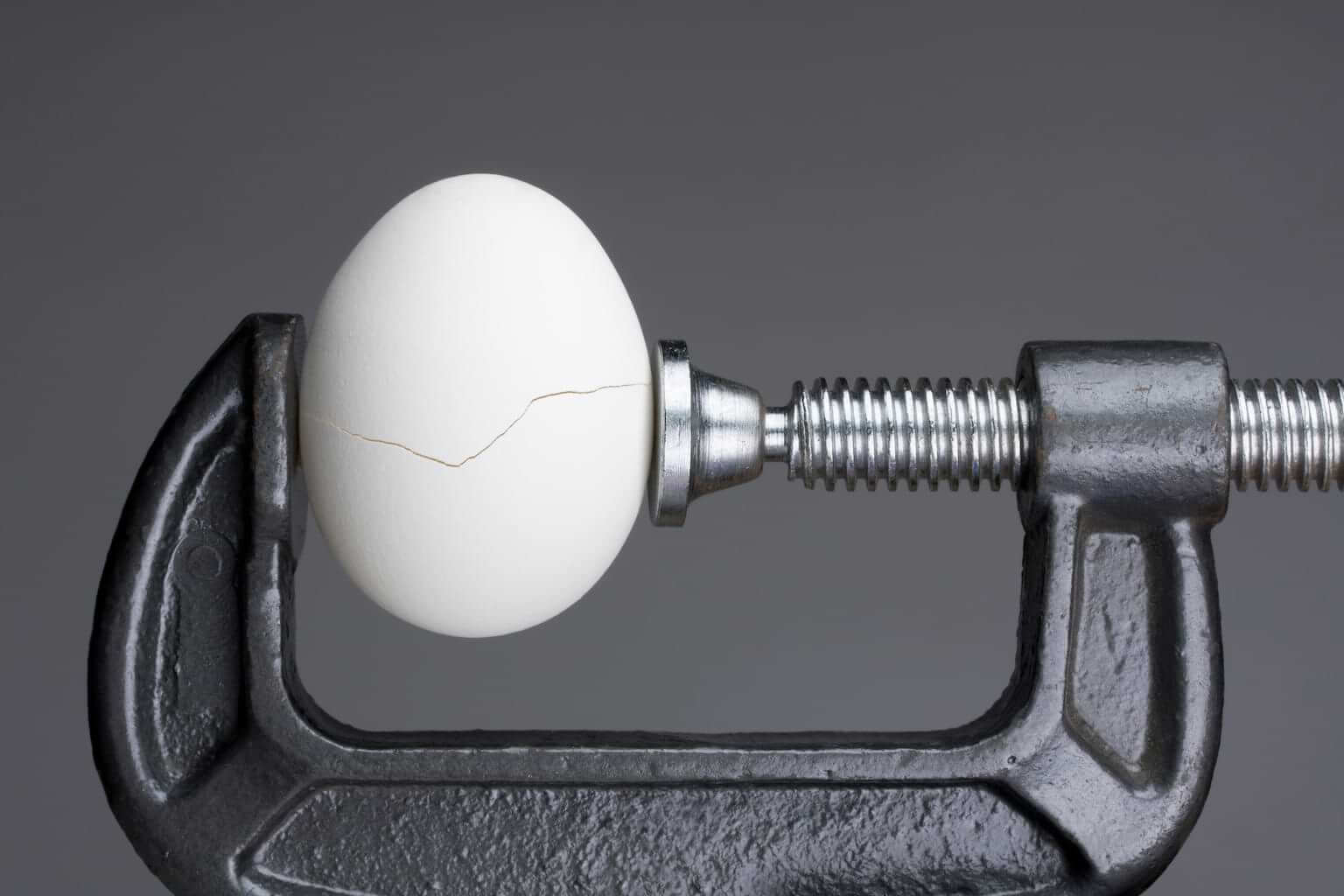 A vice is tightened around a white egg with a small crack running along its center. By killykoon/stock.adobe.com