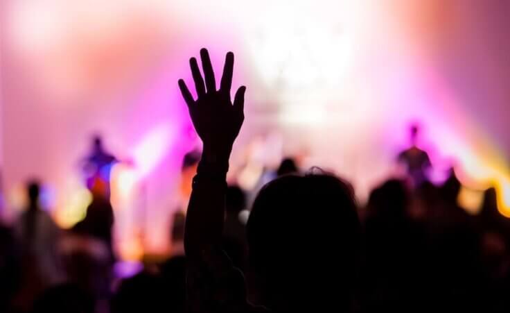 In silhouette, a woman raises her left hand into the air during a worship service. © By aradaphotography/stock.adobe.com