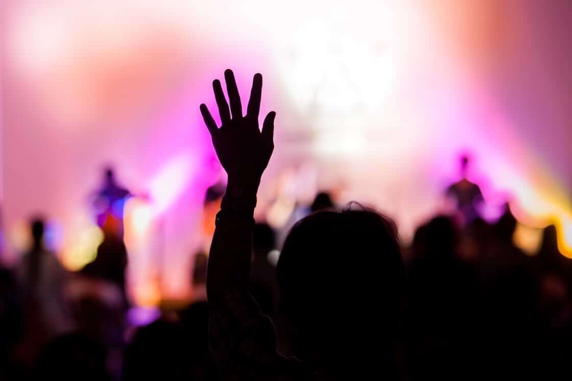 In silhouette, a woman raises her left hand into the air during a worship service. © By aradaphotography/stock.adobe.com