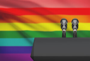 Two microphones on a pulpit in front of an LGBTQ+ rainbow flag. By niyazz/stock.adobe.com.