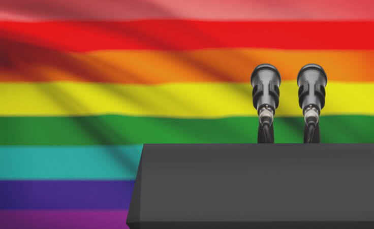Two microphones on a pulpit in front of an LGBTQ+ rainbow flag. By niyazz/stock.adobe.com.