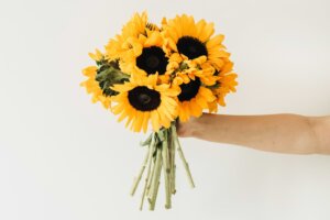 An outstretched arm holds a bouquet of sunflowers against an off-white background. By Floral Deco/stock.adobe.com.