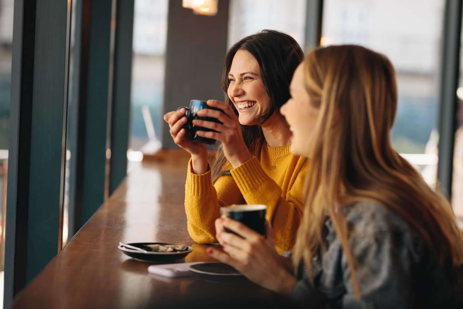 Two young women simle while enjoying coffee at a coffeeshop. By Jacob Lund/stock.adobe.com. What is the secret to happiness?