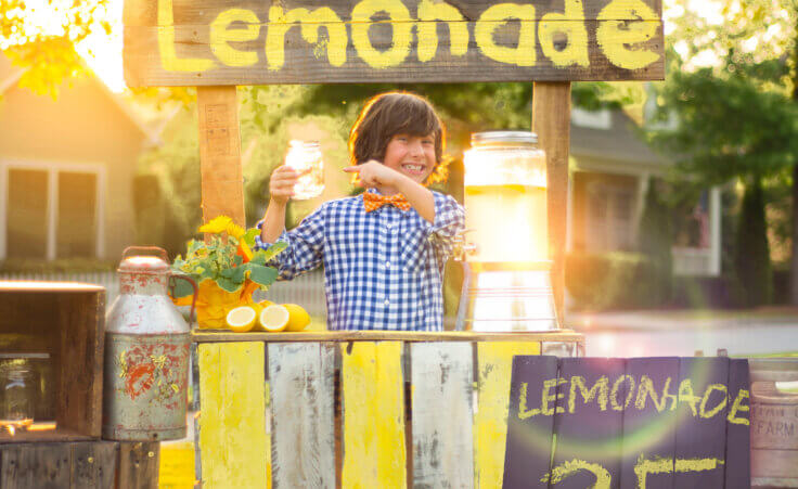 Stock photo: A boy in a blue-checkered shirt and orange bow tie stands behind his lemonade stand smiling as he points to the glass of lemonade he's holding up in his right hand. By Jim/stock.adobe.com. The photo represents Edison Juel's "Lemonade for Lahaina" lemonade stand.