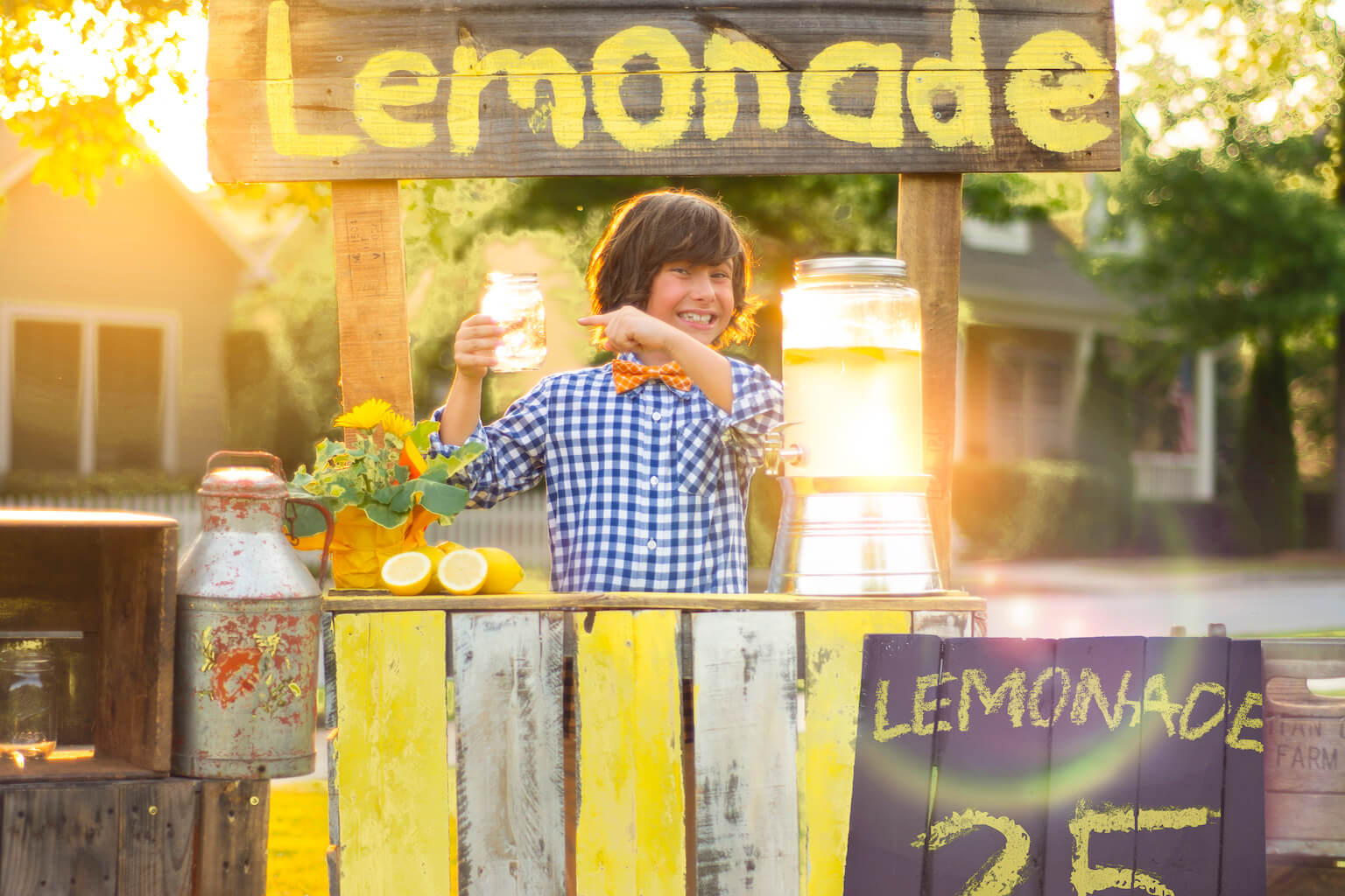 Stock photo: A boy in a blue-checkered shirt and orange bow tie stands behind his lemonade stand smiling as he points to the glass of lemonade he's holding up in his right hand. By Jim/stock.adobe.com. The photo represents Edison Juel's "Lemonade for Lahaina" lemonade stand.