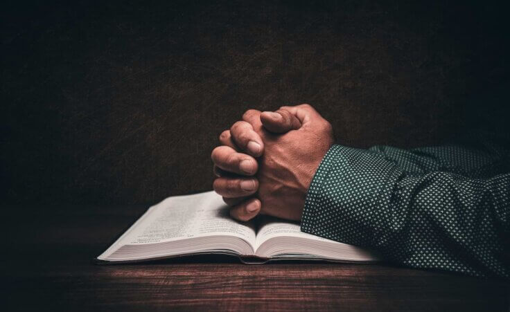 Hands folded in prayer on top of a Bible. By JavierArtPhotography/stock.adobe.com