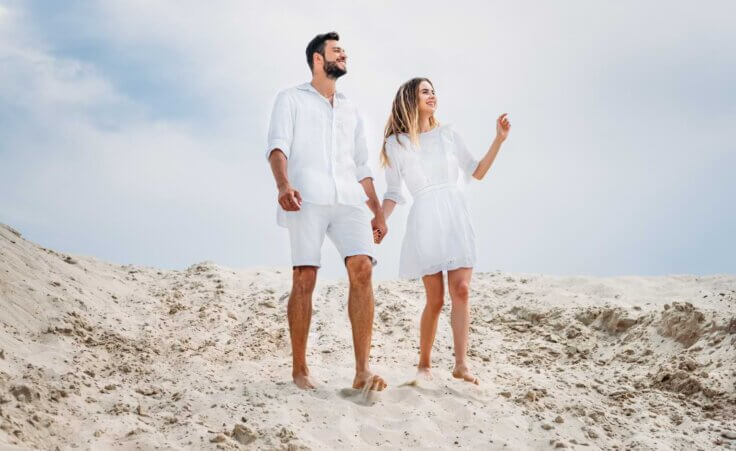 A man and woman, both dressed in white summer clothes, hold hands as they walk barefoot on a sandy dune. By LIGHTFIELD STUDIOS/stock.adobe.com. An illustration of the origin of why you can't white after Labor Day