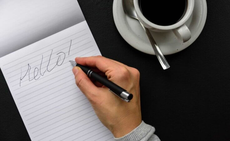 A right hand holds a pen above a notepad with the word "Hello" scribbled on it in bad handwriting. By molenira/stock.adobe.com