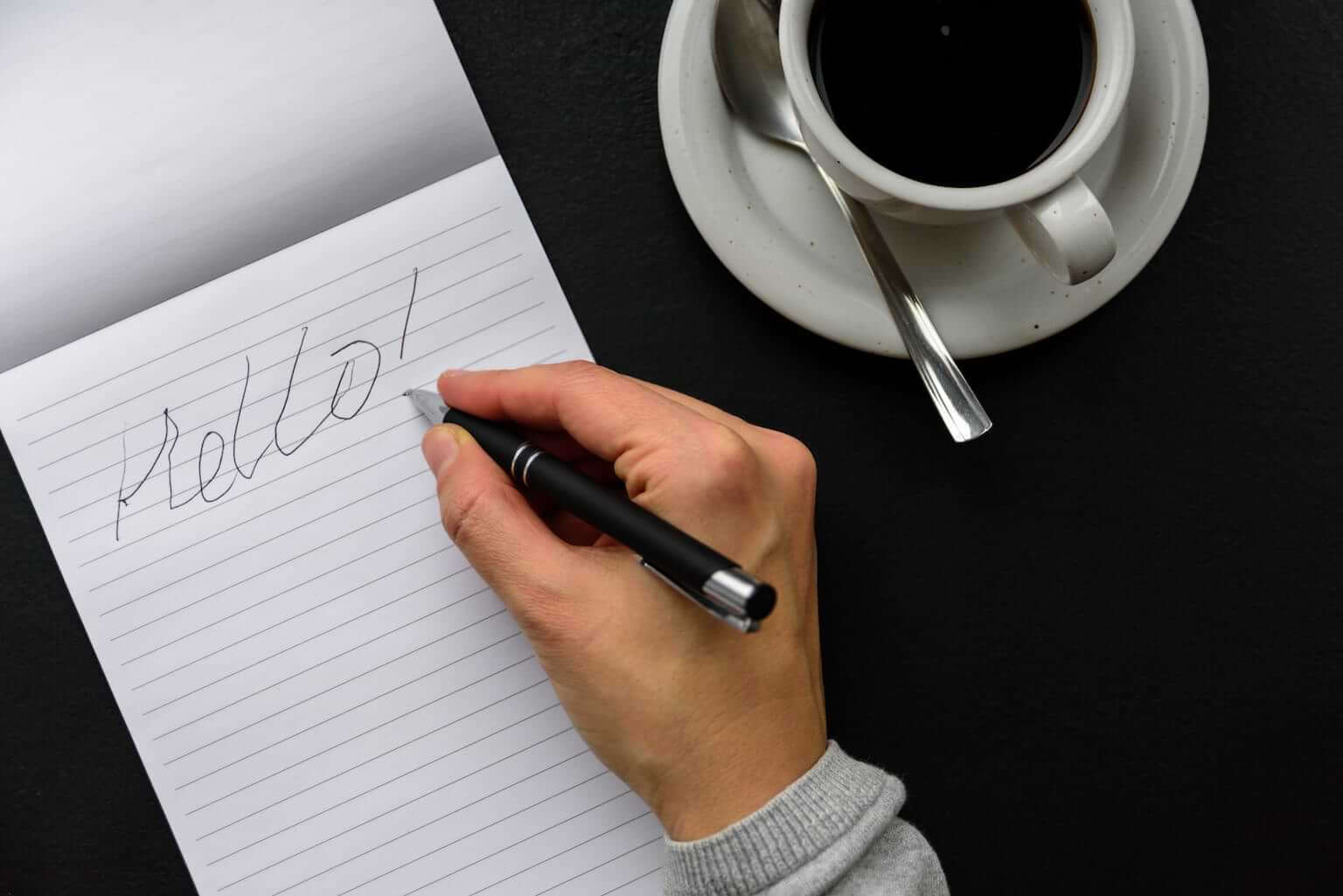 A right hand holds a pen above a notepad with the word "Hello" scribbled on it in bad handwriting. By molenira/stock.adobe.com