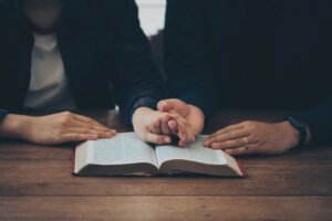 A couple holds hands in prayer over an open Bible on a wooden table. By Pcess609/stock.adobe.com