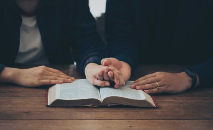 A couple holds hands in prayer over an open Bible on a wooden table. By Pcess609/stock.adobe.com