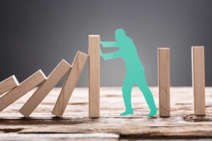 A green cutout of a man presses against leaning dominoes, preventing more from falling. By Andrey Popov/stock.adobe.com
