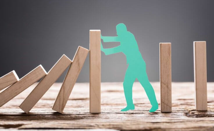 A green cutout of a man presses against leaning dominoes, preventing more from falling. By Andrey Popov/stock.adobe.com