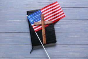 A small wooden cross sits atop an American flag which sits atop a closed Bible. By Pixel-Shot/stock.adobe.com
