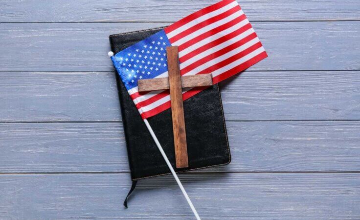 A small wooden cross sits atop an American flag which sits atop a closed Bible. By Pixel-Shot/stock.adobe.com