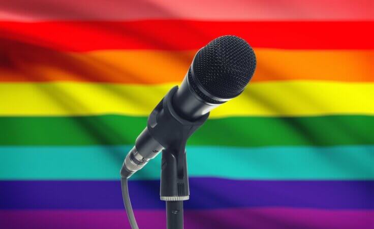 A microphone on a stand in front of a rainbow LGBTQ flag. By niyazz/stock.adobe.com