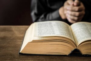 A man clasps his hands in prayer on top of a table behind an open Bible. By fotoduets/stock.adobe.com