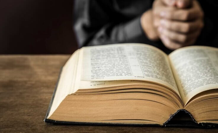 A man clasps his hands in prayer on top of a table behind an open Bible. By fotoduets/stock.adobe.com