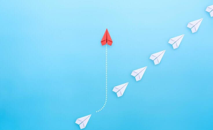 A red paper airplane diverts its path from the status quo travel of a line of white paper airplanes traveling in a straight, diagonal direction. By Worawut/stock.adobe.com