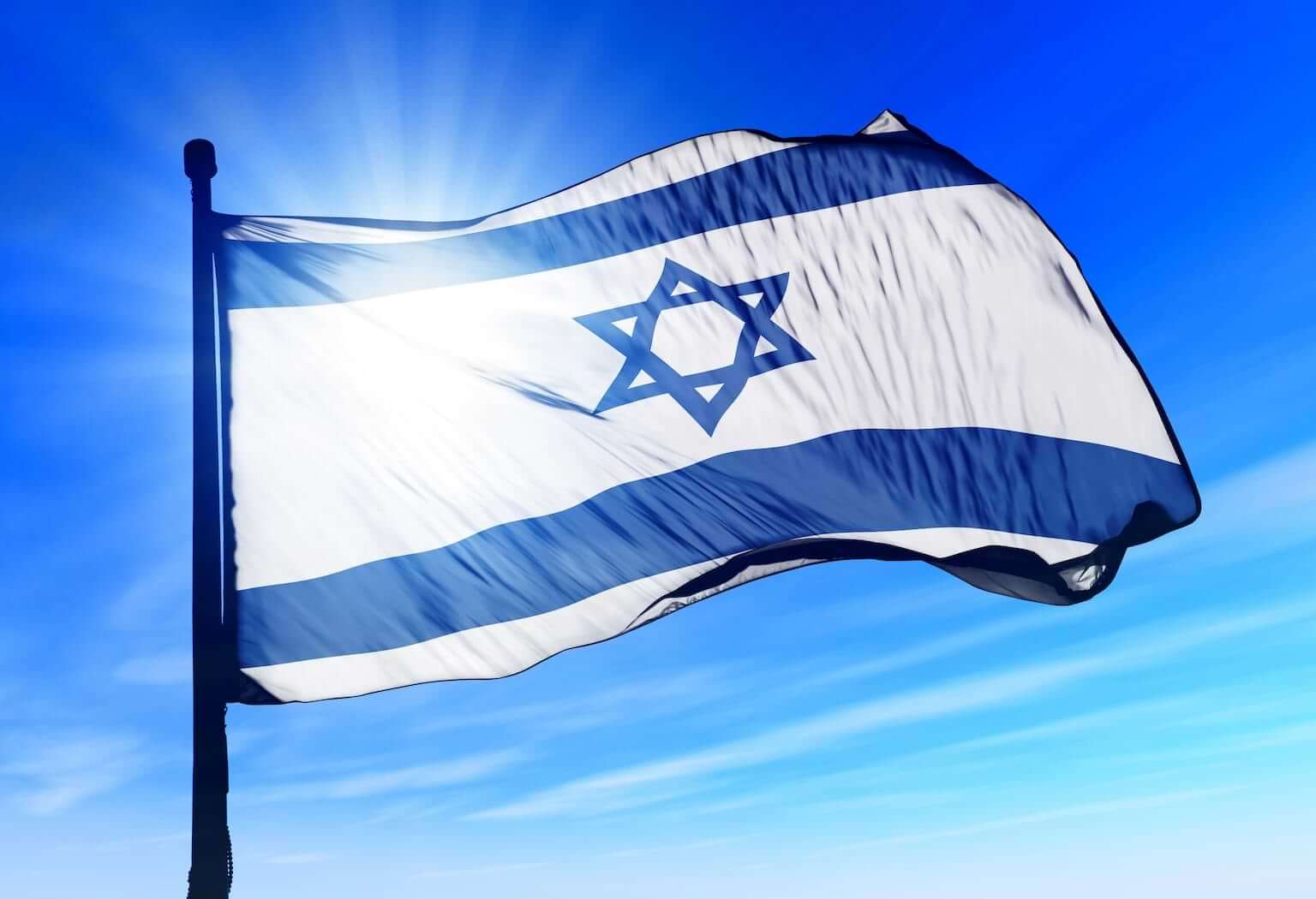 The sun shines through a waving flag of Israel. By Lulla/stock.adobe.com