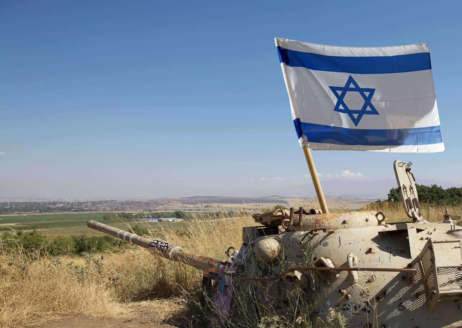 An Israeli tank sits in a field with an Israel flag upright within the tank. By Maurizio/stock.adobe.com