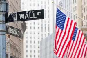 Street signs for Wall Street and Broad Street in New York City stand in front of two large American flags hanging from the side of a skyscraper. By kasto/stock.adobe.com